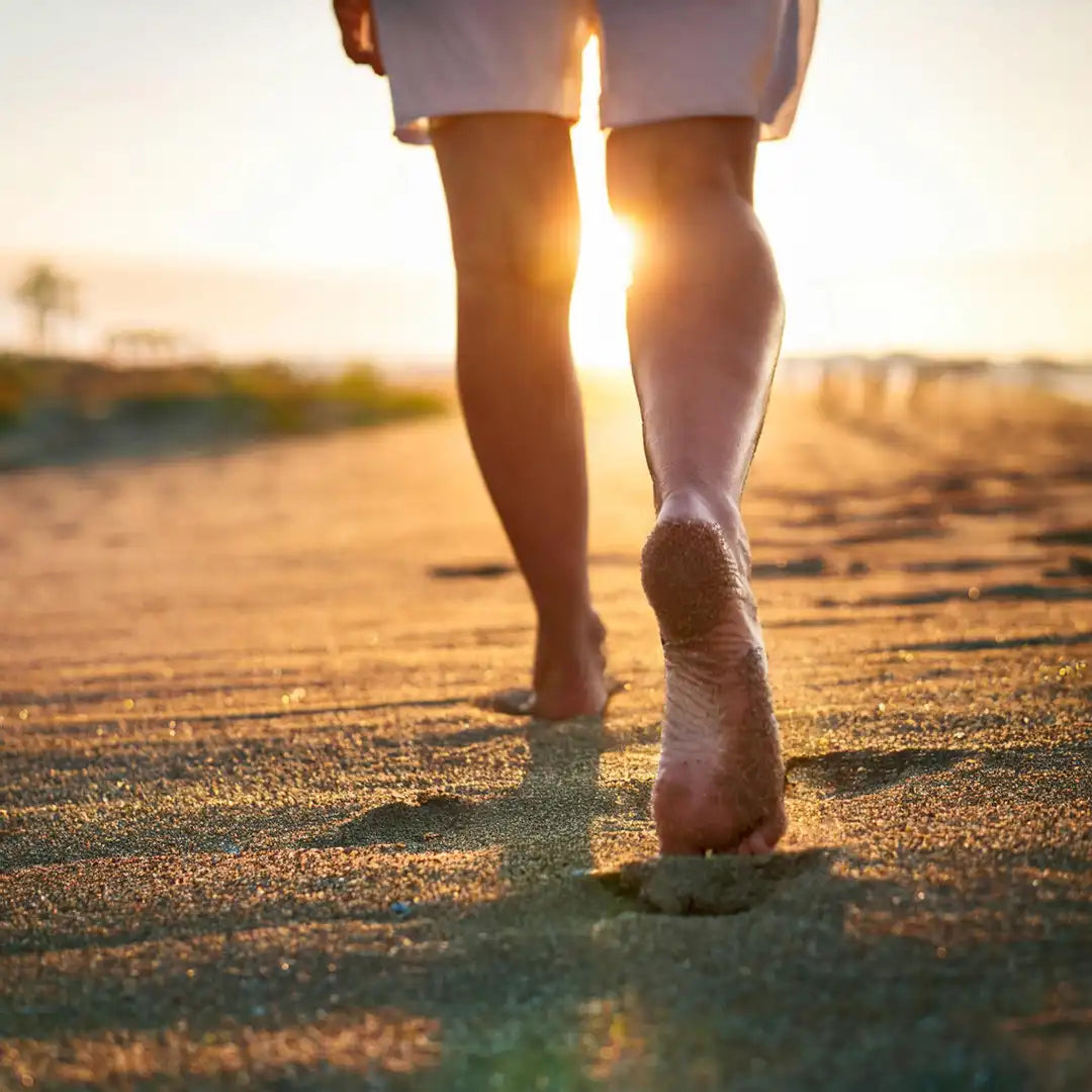 Eine Person geht barfuß am Strand entlang, während die Sonne untergeht. Natürliches Gehen auf Sand fördert die Fußgesundheit und entspannt die Muskulatur.